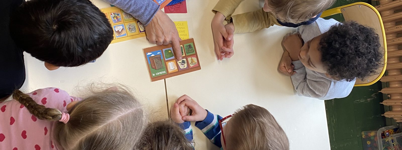 Vue aérienne de sept enfants et d'un adulte participant à un jeu éducatif haut en couleur autour d'une table. L'adulte montre du doigt des cartes de jeu, tandis que les enfants l'observent attentivement.