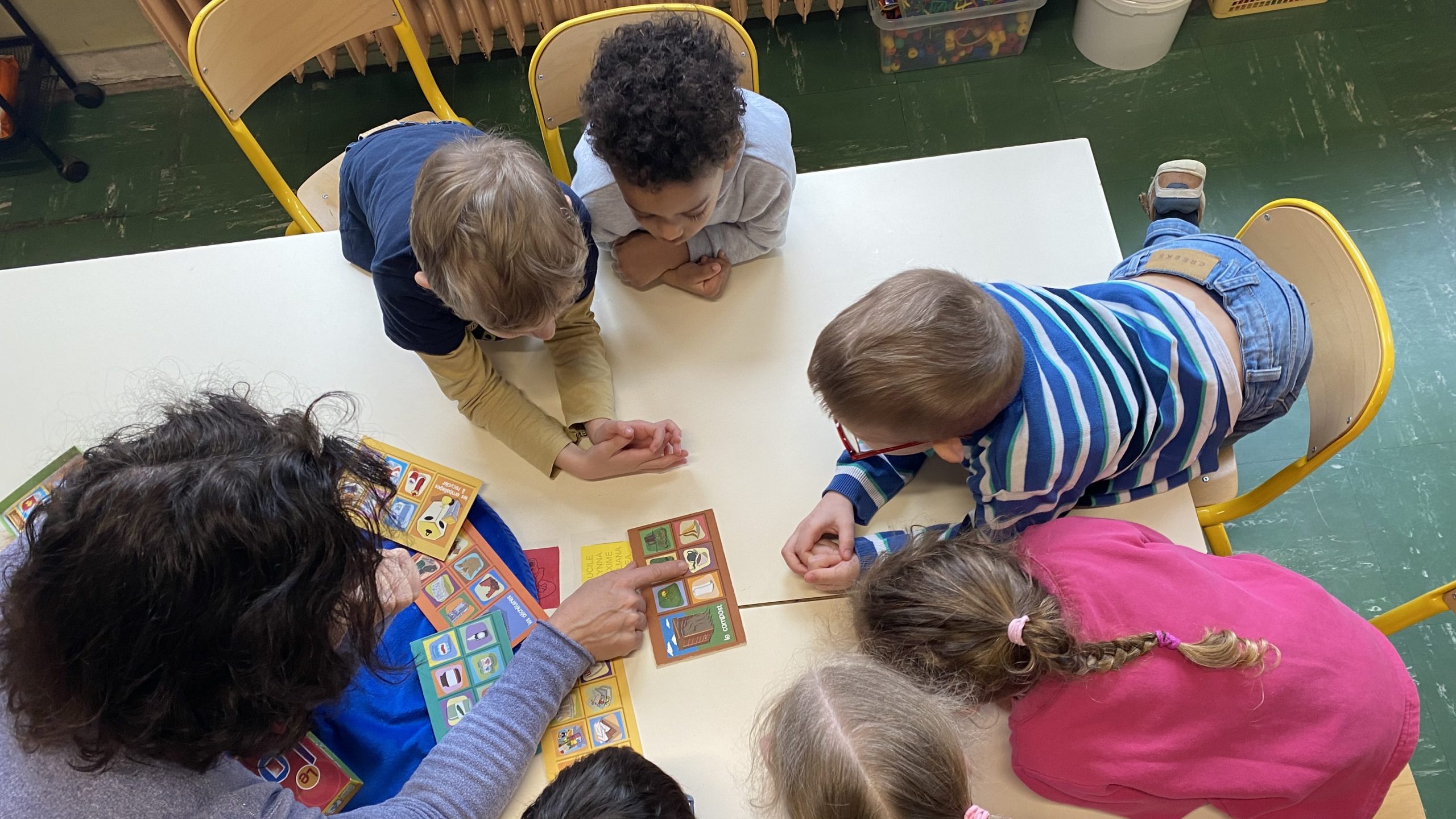 Vue aérienne de sept enfants et d'un adulte participant à un jeu éducatif haut en couleur autour d'une table. L'adulte montre du doigt des cartes de jeu, tandis que les enfants l'observent attentivement.